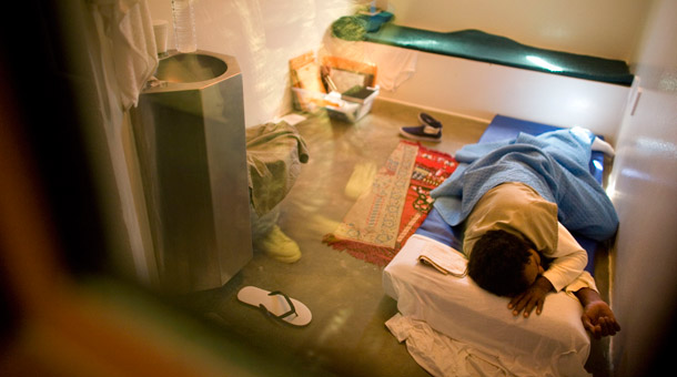 In this image reviewed by the U.S. Military, a Guantanamo detainee, photographed through a glass window with a U.S military guard seen reflected on it, sleeps on a mattress on the floor of his cell, at the Camp 5 detention facility, at Guantanamo Bay, Cuba. (AP Photo/Brennan Linsley)