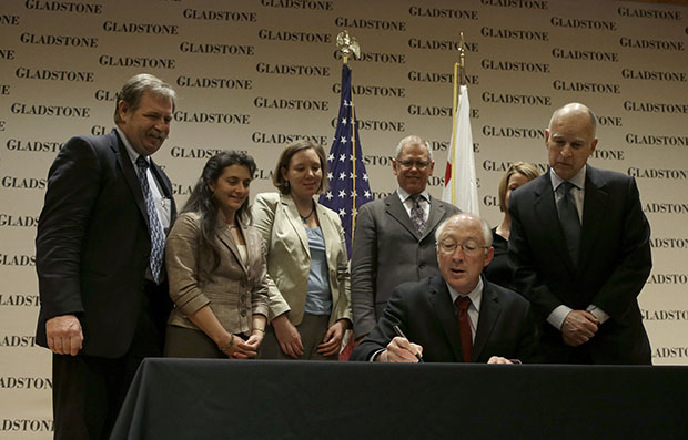 Former U.S. Secretary of the Interior Ken Salazar, bottom, signs a bill in front of Gov. Jerry Brown (D-CA), right, and others after a news conference at the Gladstone Institutes in San Francisco, California, Wednesday, March 13, 2013. (AP/Jeff Chiu)