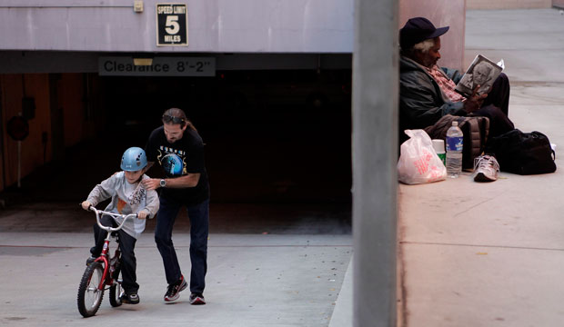 David Row helps his 8-year-old son Lestat learn to ride a bicycle as a homeless man reads outside a homeless shelter in Los Angeles on September 14, 2011. (AP/Jae C. Hong)