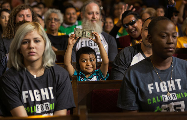 A child, center, with his father, right, raises a dollar bill as workers await the Los Angeles City Council's vote to raise the city's minimum wage to $15 per hour by 2020, June 3, 2015. (AP/Damian Dovarganes)