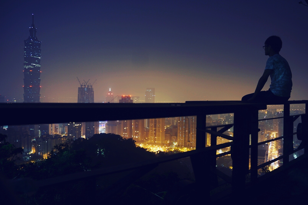 A man sitting on a balcony looks out over Taipei at night.