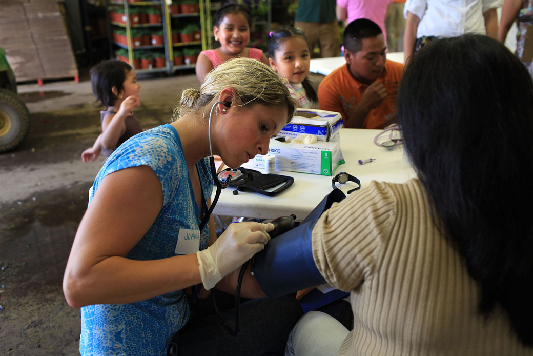  (A college students takes a woman's pulse at a flower nursery serving as a health care clinic for migrant farmworkers in Connecticut.)