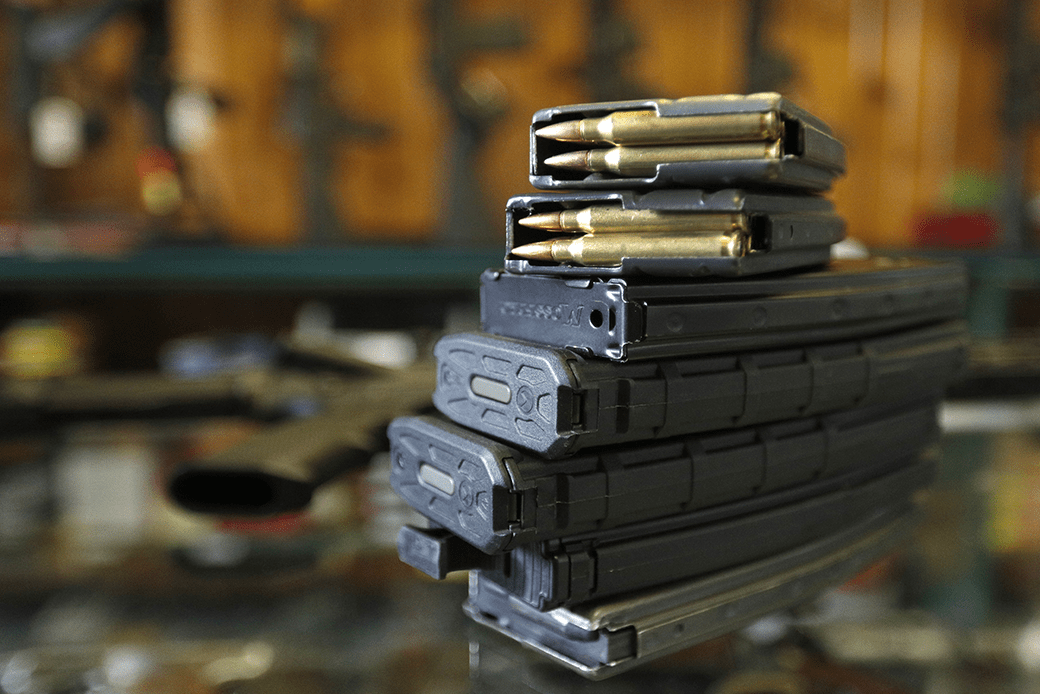  (A stack of 30-round and other high-capacity clips for use in an AR-15 is pictured on a table surface in a shooting range.)