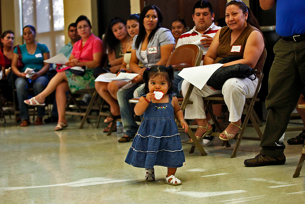  (A one-year-old girl is shown standing and eating a cookie, surrounded by Latinx parents attending a school board meeting.)