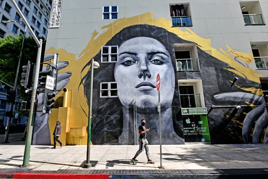 A man wearing a face mask walks past a mural of a woman on a street that is otherwise empty due to shelter-in-place orders in response to the COVID-19 outbreak in Los Angeles, March 2020. (Getty/Michael Ochs Archives/Stephen Albanese)