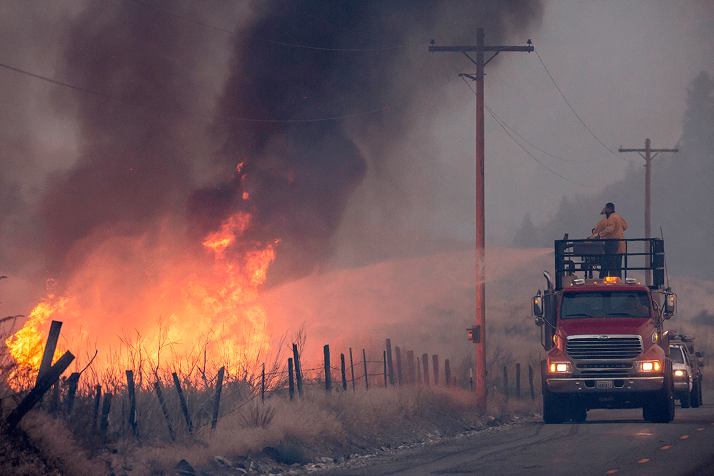  (A man on top of a makeshift firetruck sprays water onto a wildfire.)