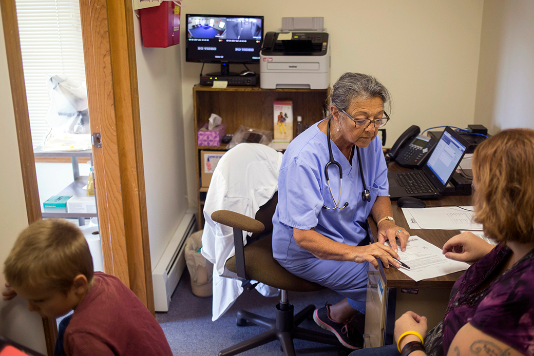  (A nurse practitioner sits at her desk across from a patient and goes over details on a sheet of paper; the patient's young son is partially seen in the bottom left corner.)