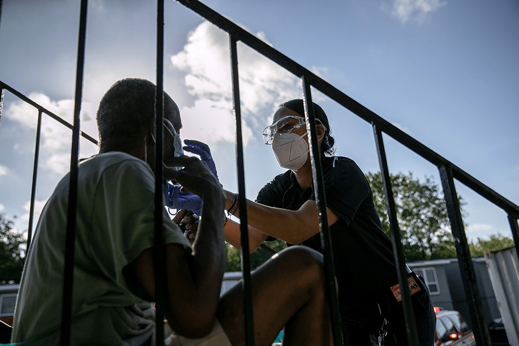 (A masked female medic with the fire department is shown providing oxygen to a seated man who is having trouble breathing.)