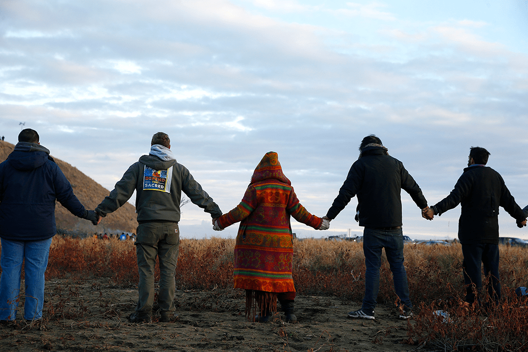  (A row of five people (part of a longer chain) holding hands, backs to the viewer. They are standing in a field looking out to a blue/gray sky, seemingly in the evening, with police barely visible in the distance.)
