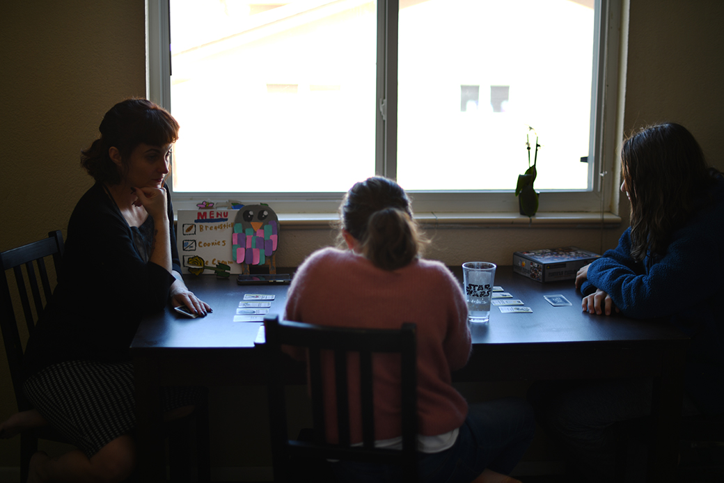  (A mother and her two children are seated inside at a table playing a card game in front of a large window.)