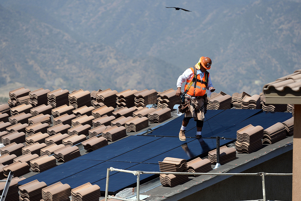 (A worker in a construction vest and hard hat installs solar panels on a roof in California, with mountains pictured in the background.)