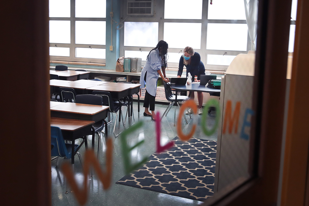  (Two female teachers are pictured in an empty classroom talking and bent forward looking at a laptop screen; picture is taken from the other side of a door with a glass pane with a 