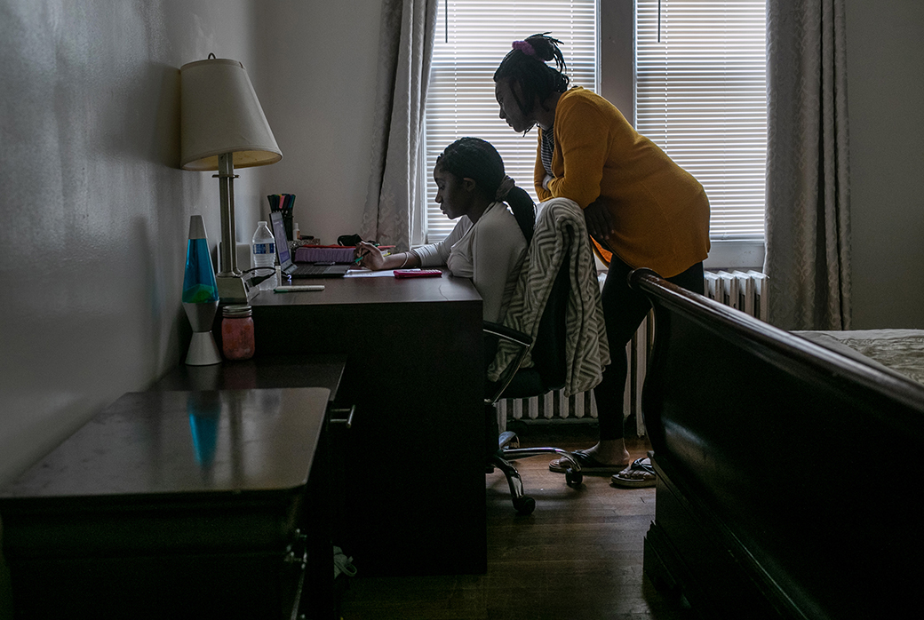 A teenage girl sits at a desk to take part in remote distance learning on a laptop, while her mother stands behind