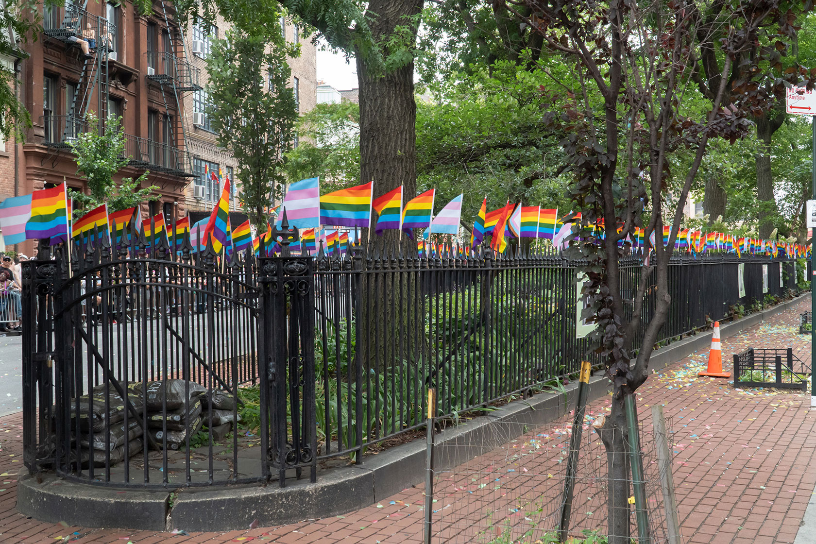 Photo shows a black wrought iron fence next to a brick sidewalk. Small, colorful pride and transgender flags line the fence.