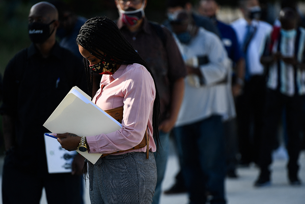 In focus, a black woman looking down and wearing a light pink sweater holds a manila folder. Behind her is a line of people wearing masks.