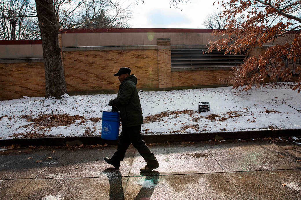 View of a Black man in profile, wearing dark winter clothes, walking on a sidewalk holding a large blue bucket. He is scattering de-icing salt in front of a brown brick/stone building with a small grassy area between the building and the sidewalk, although it's entirely covered in a tin layer of snow with brown leaves visible.