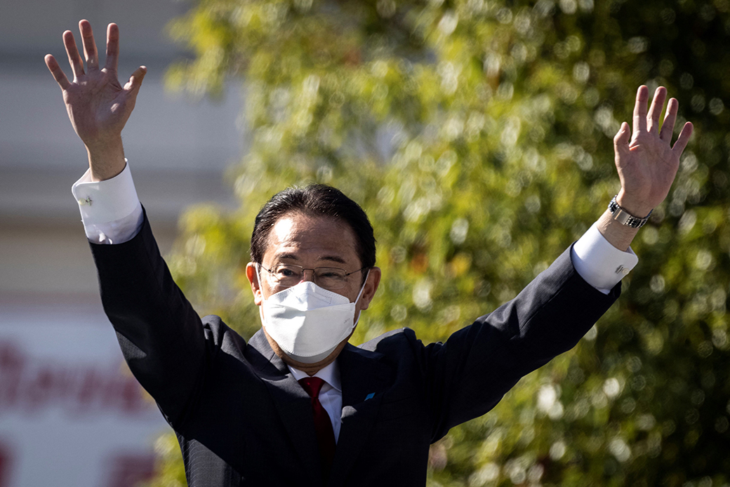 A middle aged to older Japanese man wearing a dark suit, wire glasses, and white face mask is pictured in focus with both arms raised in front of a blurred background.