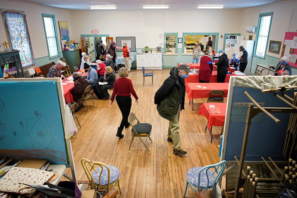 View looking into a brightly lit recreation room in a church. Folding tables covered in red tablecloths and folding chairs are visible, with several people wearing masks and winter clothes are shown sitting or standing waiting for vaccinations or filling out paper forms.