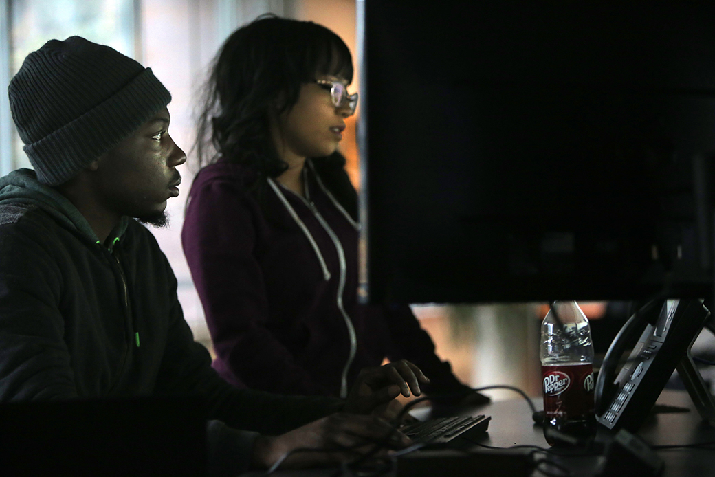 A black man wearing a dark gray beanie and a woman with dark hair and glasses, both in their twenties or thirties, are pictured staring at a large computer monitor (screen is not visible to the viewer.)