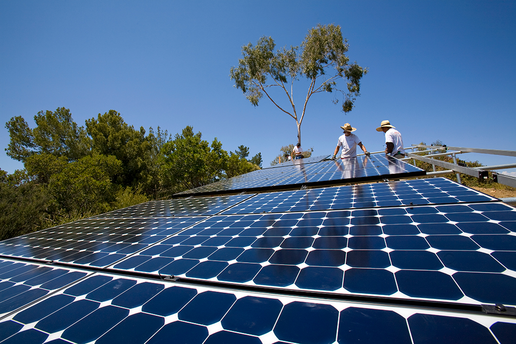 Two workers are pictured installing deep midnight blue-colored solar panels on a hill in California. Blue sky and scattered trees are seen in the background.