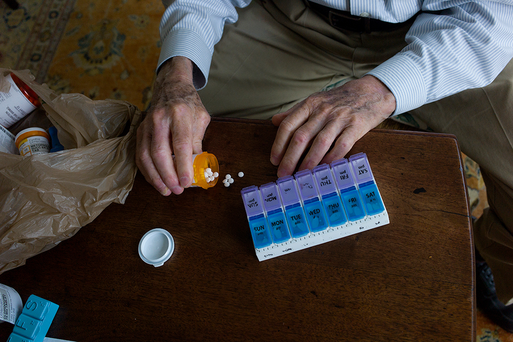 View from above of an older man's hands arranging/prepping his medication (small round white tablets from an orange Rx bottle) into a pill organizer on a brown wood coffee table. A plastic grocery bag with other bottles of prescription medication is visible on the left.