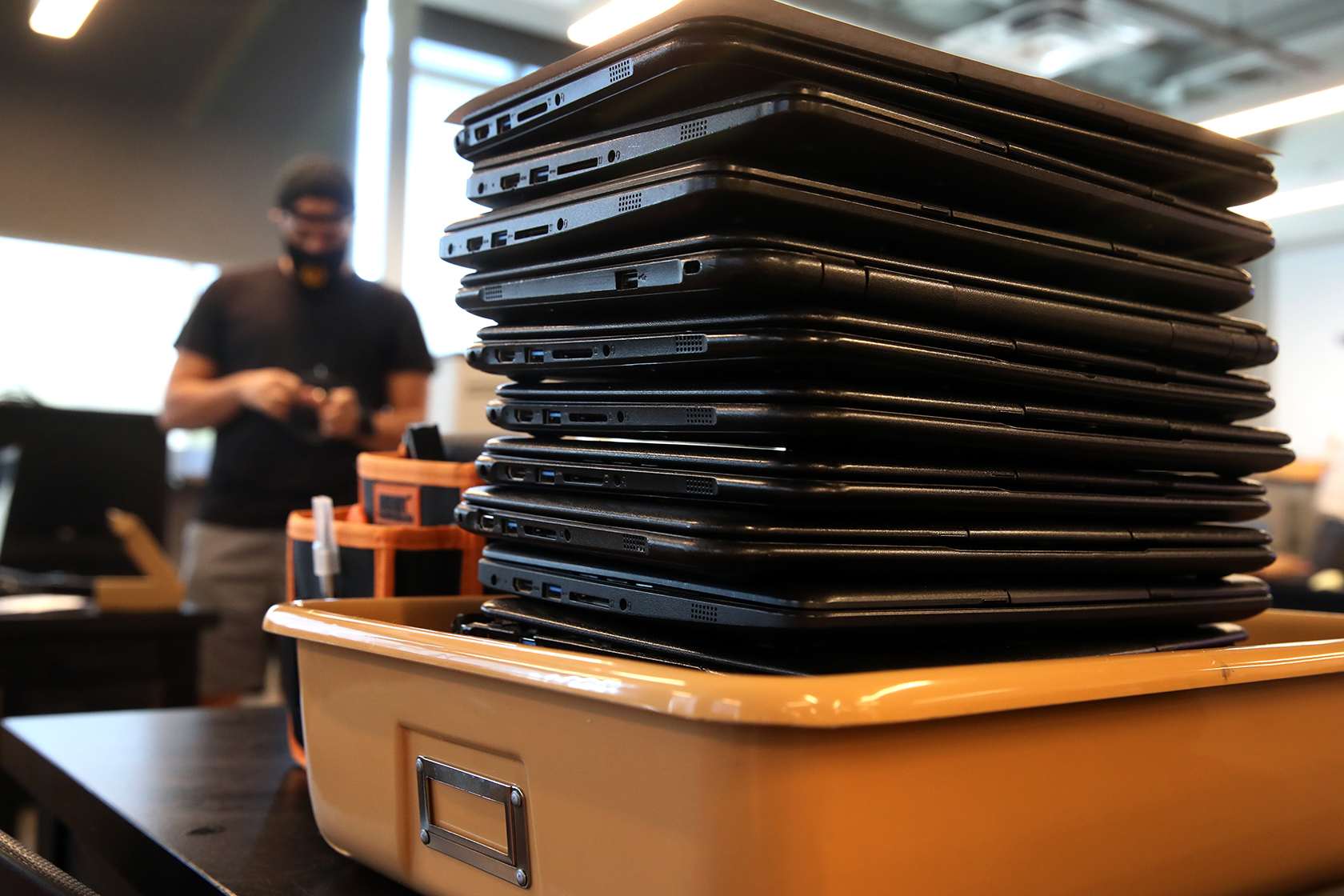 Close-up shot of a stack of black laptops in a peach-colored metal bin sitting on top of a black desk or table. Out of focus in the background, a man in a black t-shirt is pictured.