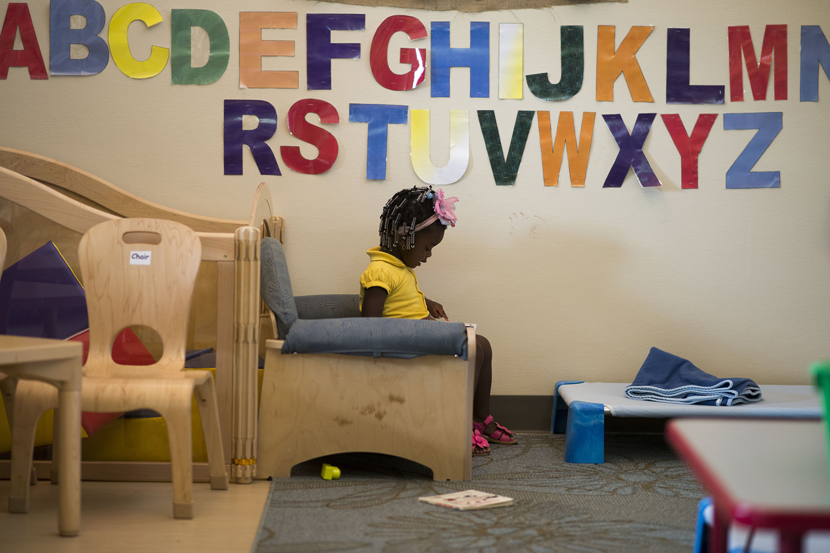 Young girl sitting in chair reading in front of alphabet letters on wall