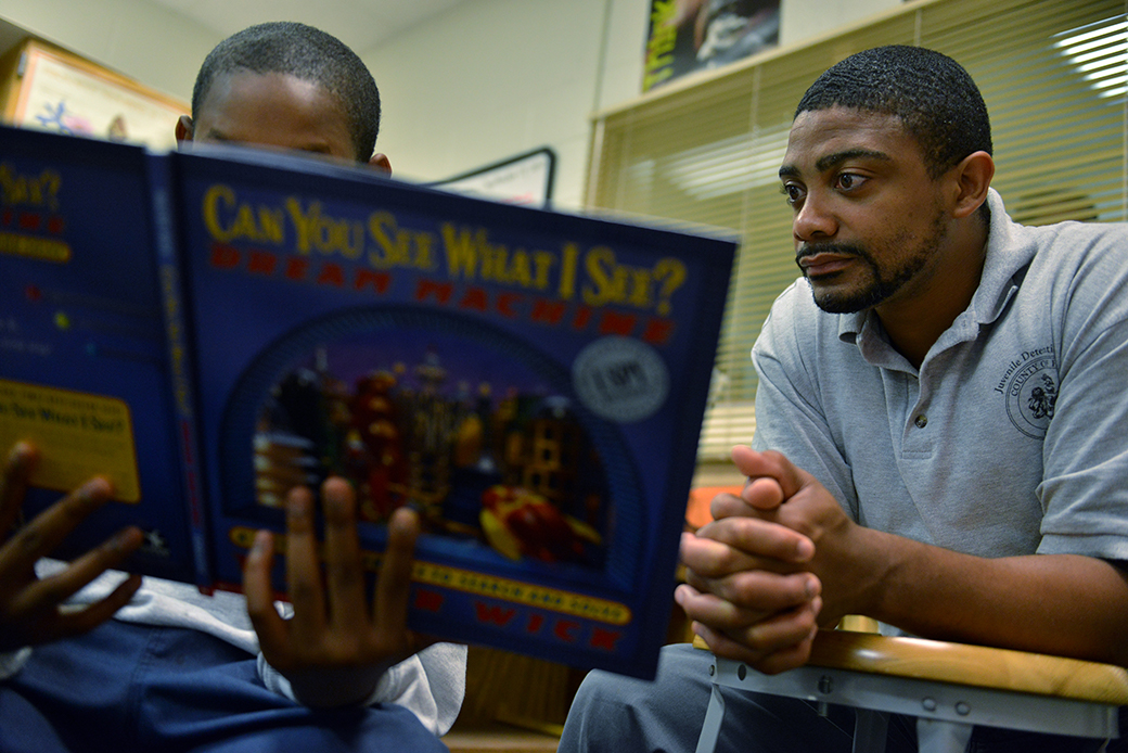A Black man wearing a gray polo shirt sits with a student (Black boy, whose face is hidden by a blue hardcover picture book)