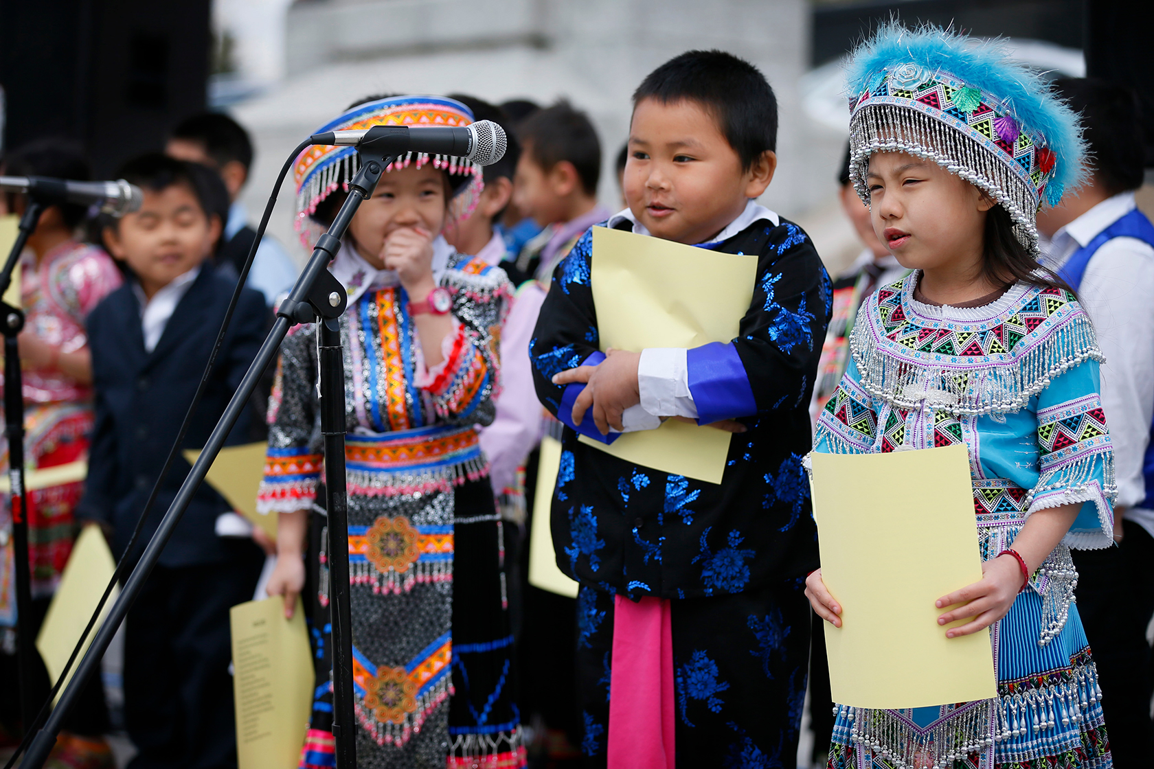A group of young (1st grade) Asian students dressed in colorful traditional clothing, standing in front of a microphone. They are holding sheets of light yellow paper, performing folk poetry for the Hmong American Day celebration at the state capitol.