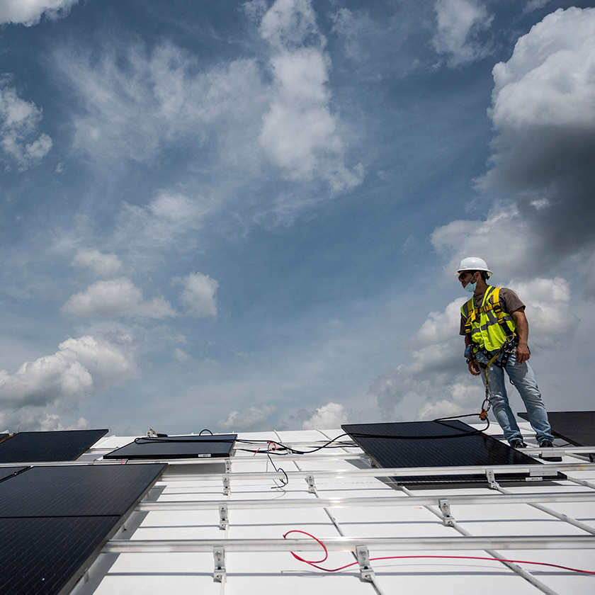 Worker wearing a bright yellow/neon visibility vest and a white protective helmet is standing on a white roof in profile, looking to the left side of the image. The background behind him is a blue sky with white clouds. At his feet is a solar panel in the process of being installed, along with some trailing wires.