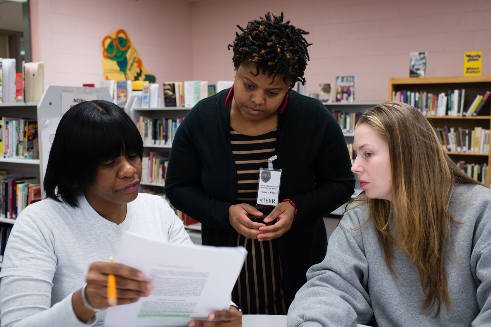 Two students and a college professor gather in a library and look at an article held by one of the women.