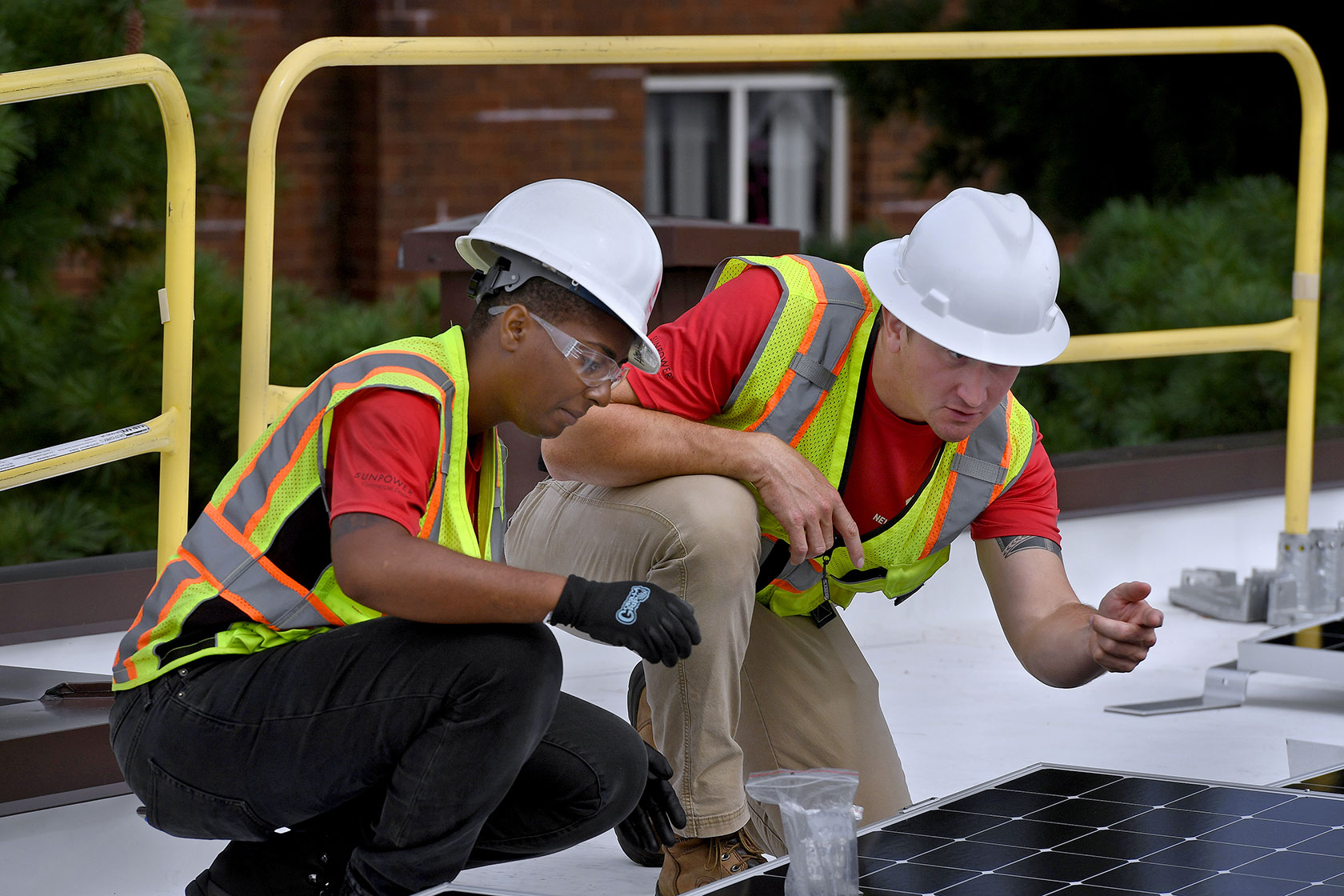 Photo shows two construction workers kneeling over a solar panel on a roof.
