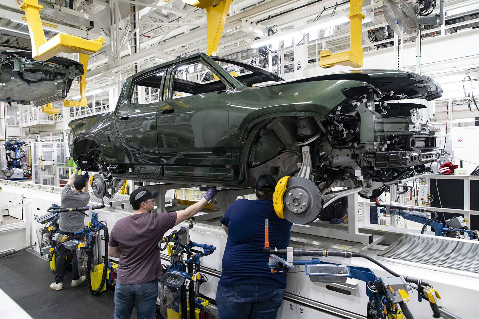 Photo shows three auto assembly workers working on the underside of a suspended electric truck.