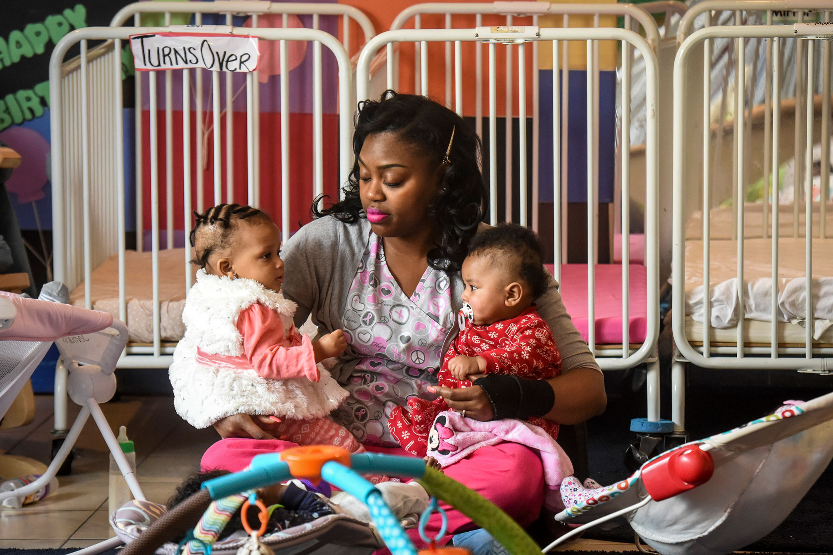 Photo shows a woman seated in front of a crib, holding an infant in each arm.