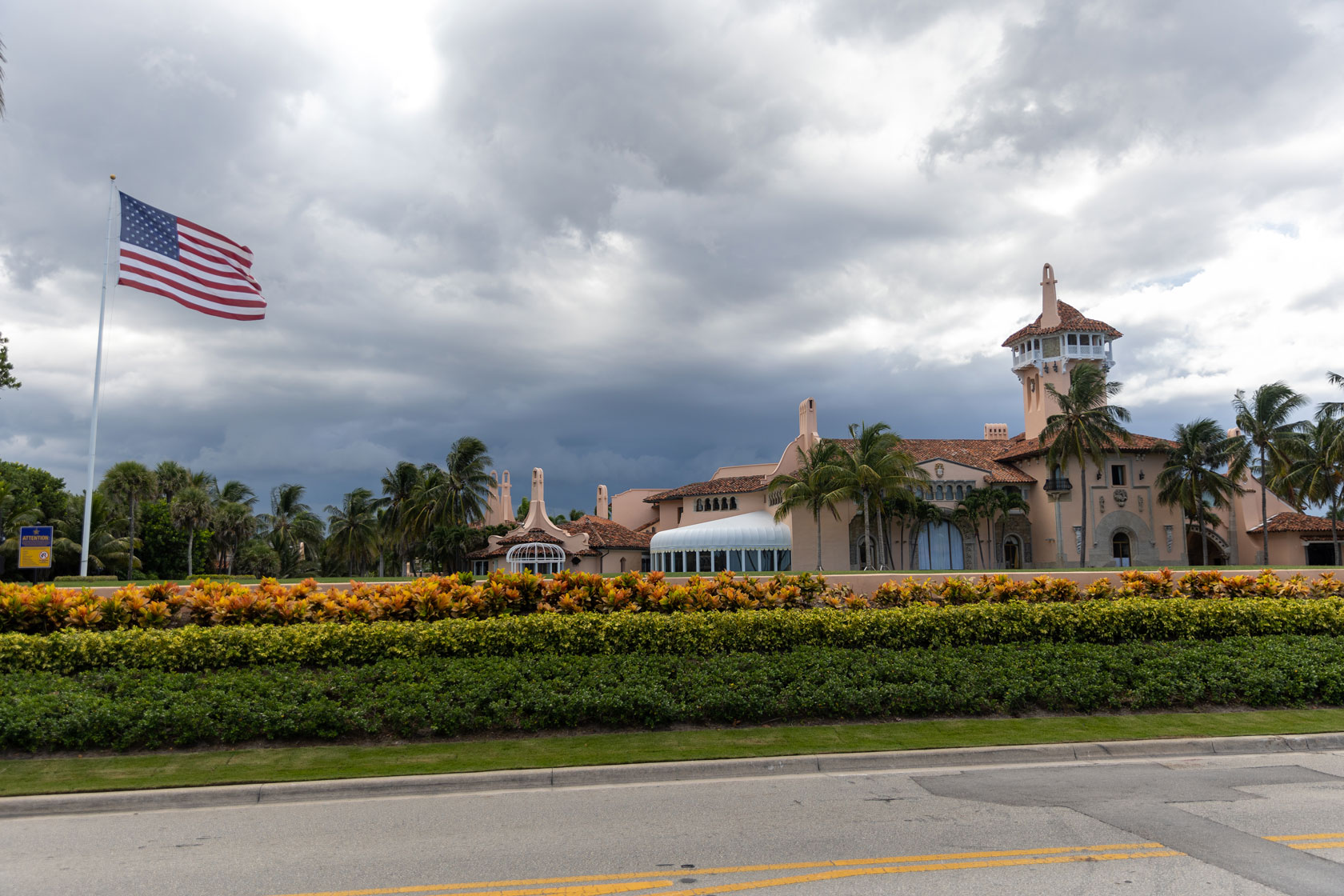 The Mar-a-Lago resort is seen against a stormy gray cloud with an American flag in front.