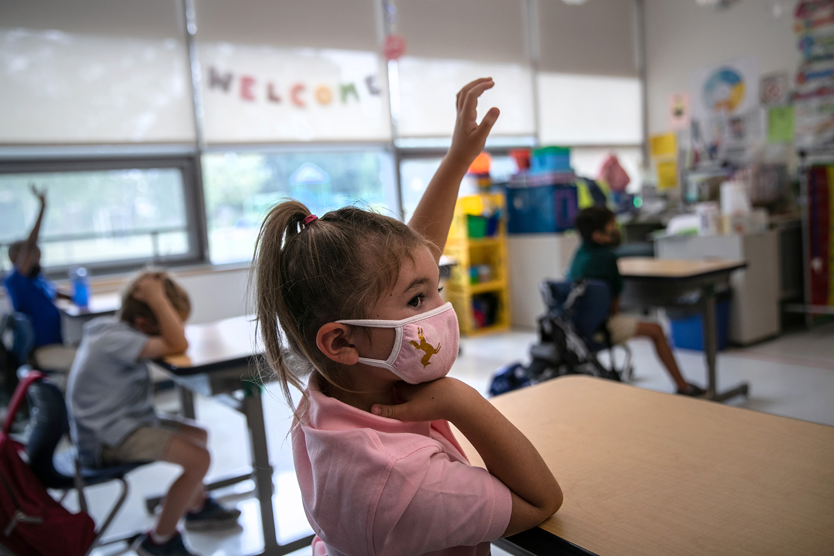 Young girl wearing face mask with other students, windows, and Welcome sign in background