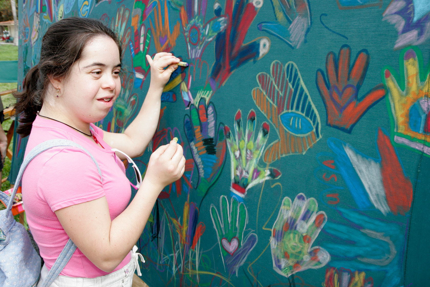 Photo shows a young woman drawing on a chalkboard covered with colorful handprints.
