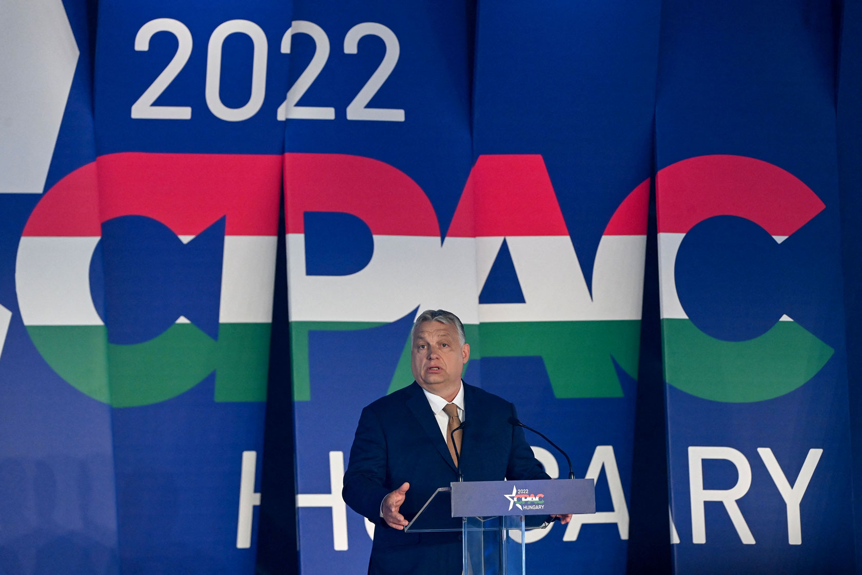 Photo shows Orbán standing at a podium in front of a large banner that says 