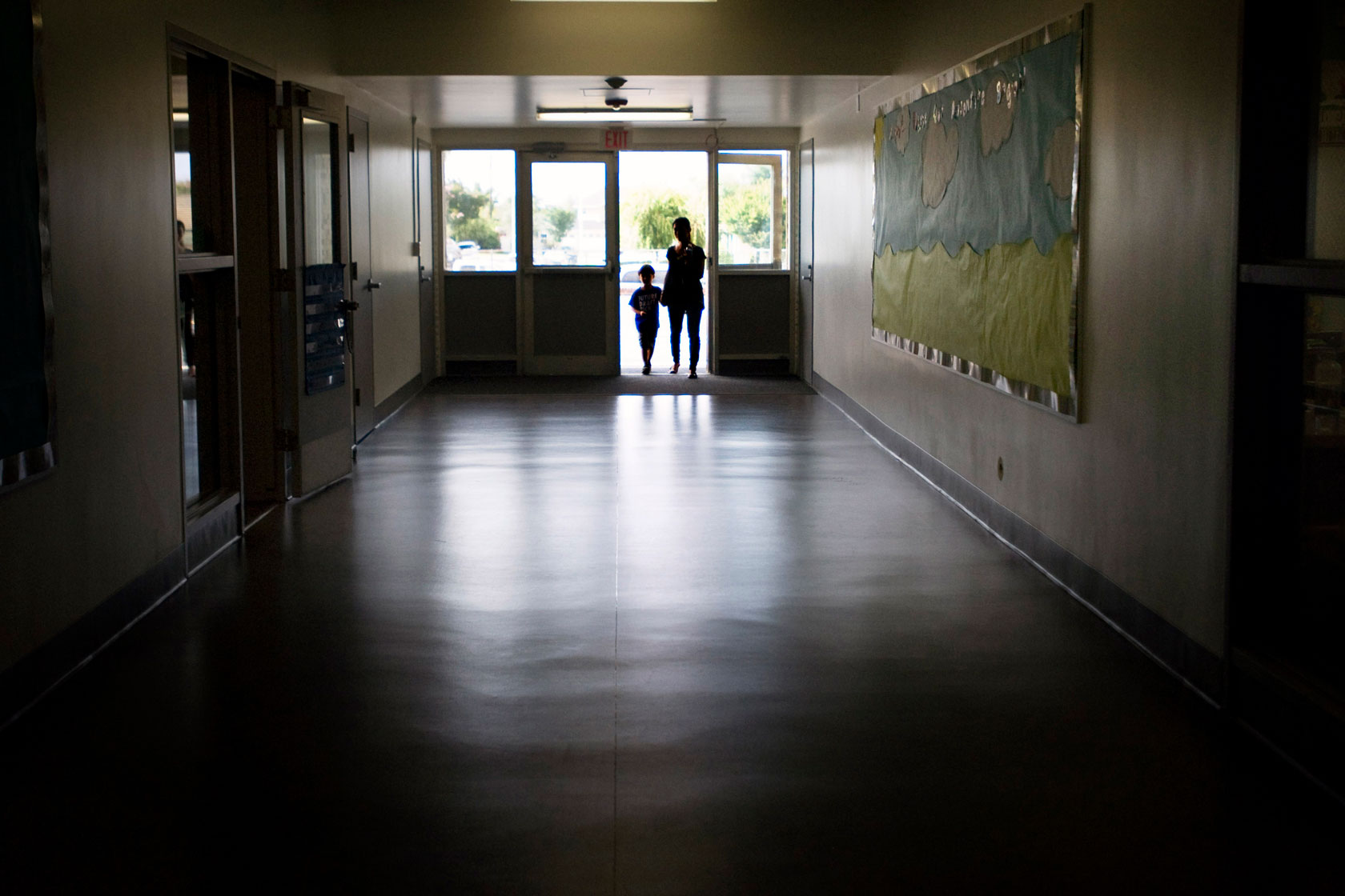 Photo shows a woman and her young son entering a doorway at the end of a dark school hallway.