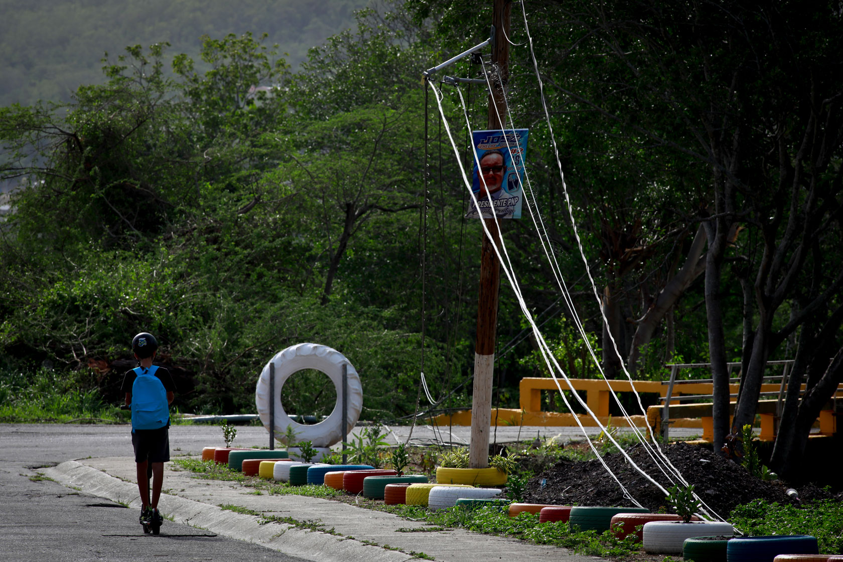 Photo shows a young child riding a scooter on a road with green trees in the background and downed power lines.