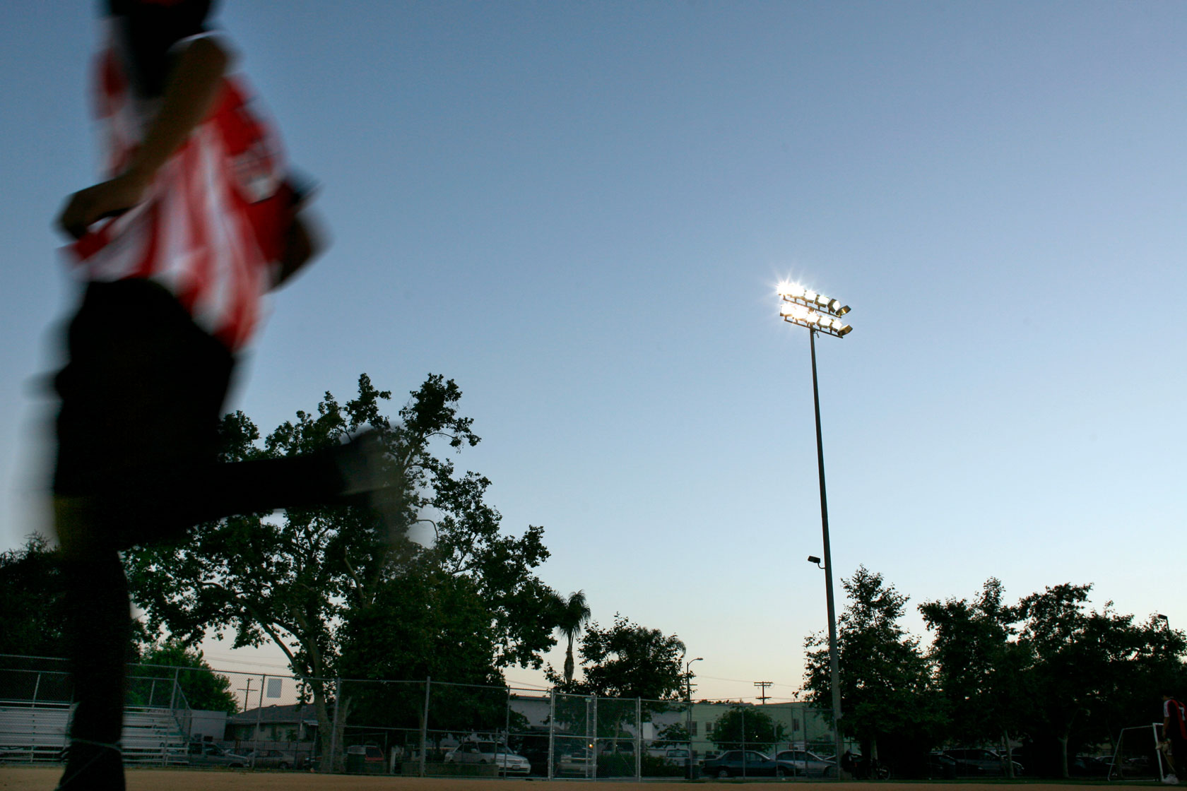 A boy runs by on a lit-up baseball field in the forefront at dusk.