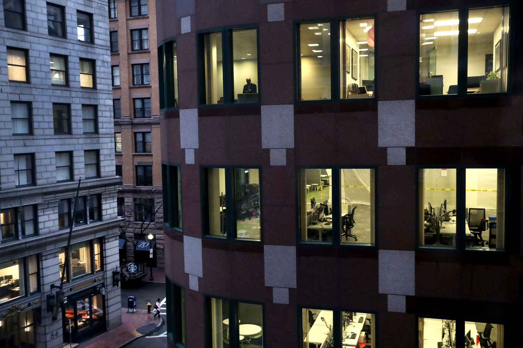 Photo shows a nearly empty office building with windows illuminated and one or two people present inside.
