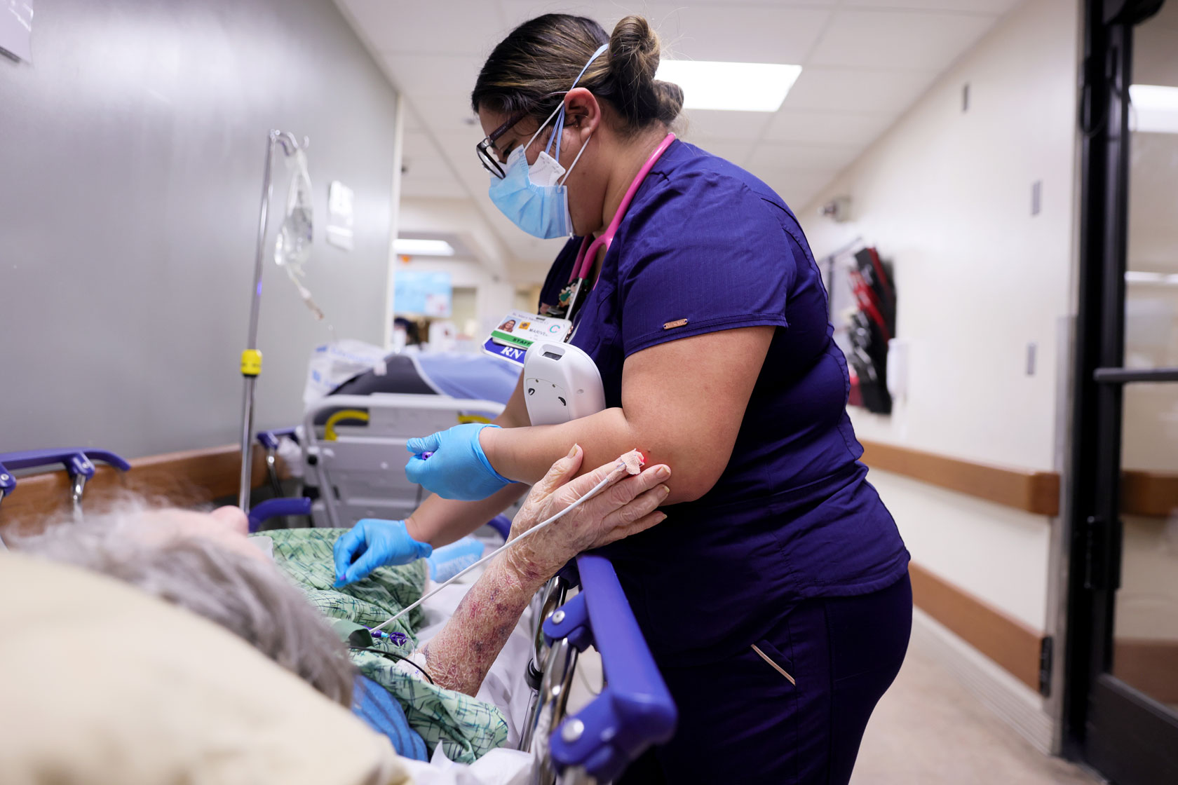 Photo shows a nurse in a purple shirt caring for a patient, who touches the nurse's arm with her hand.
