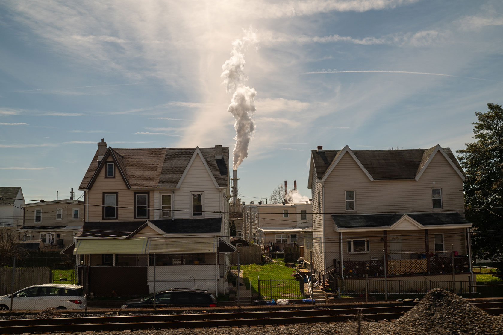 Photo shows two houses next to a train track, and a smoke stack in the background emitting a large plume of smoke.