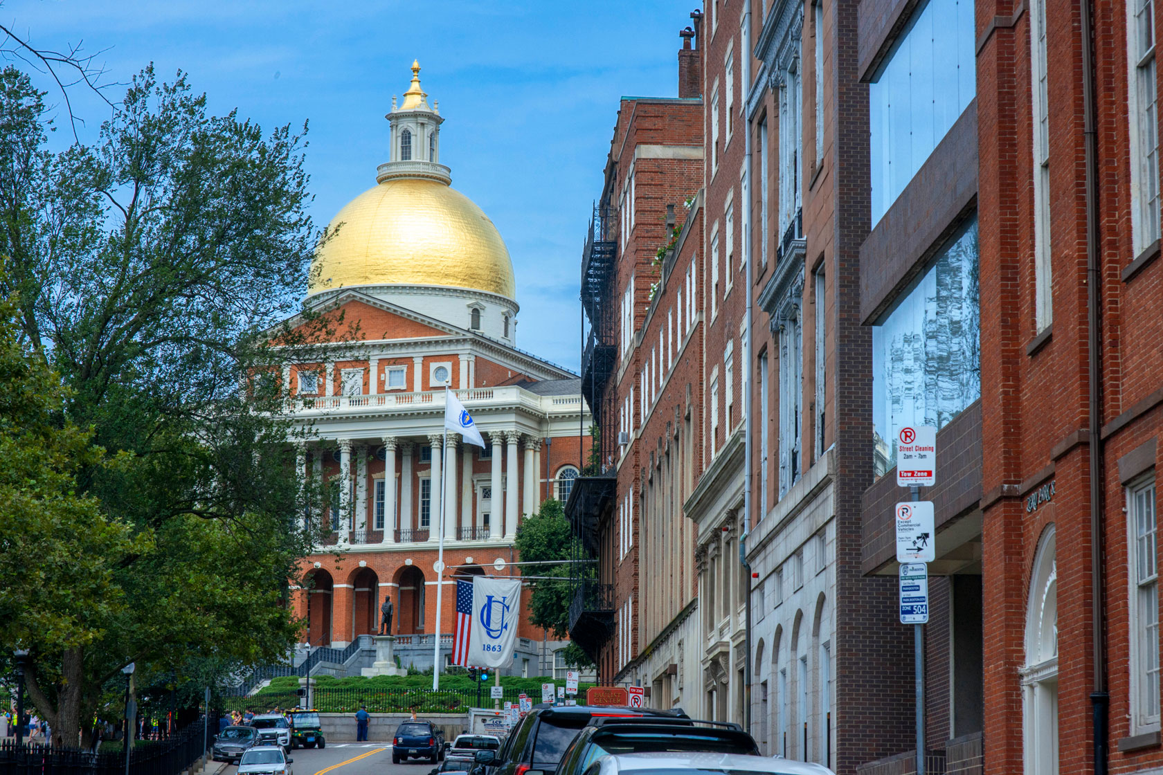 The golden dome of the Massachusetts State House is seen behind a street beneath a blue sky.