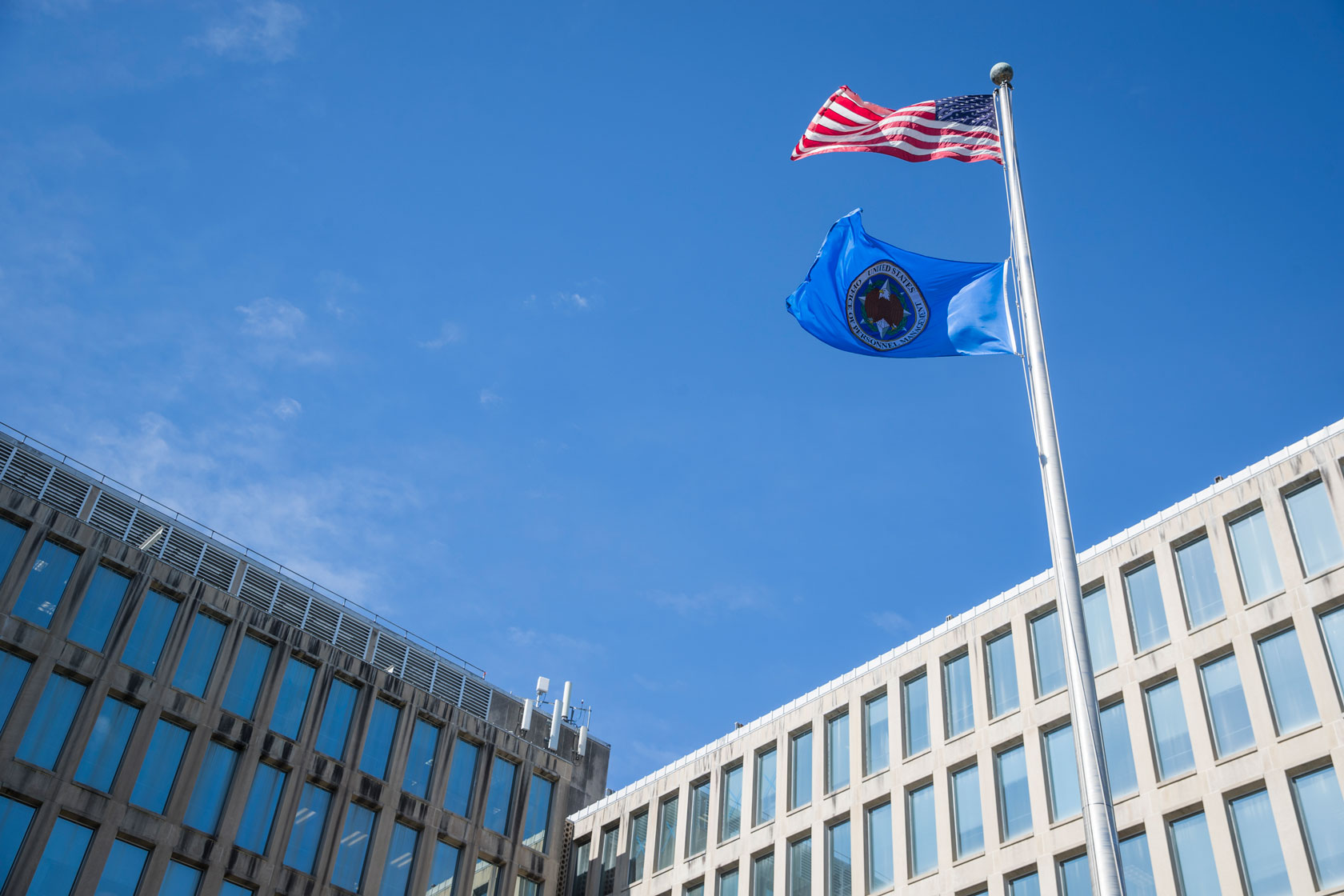 The American and U.S. Office of Personnel Management (OPM) flags blowing in the wind on a sunny day in front of the OPM building.