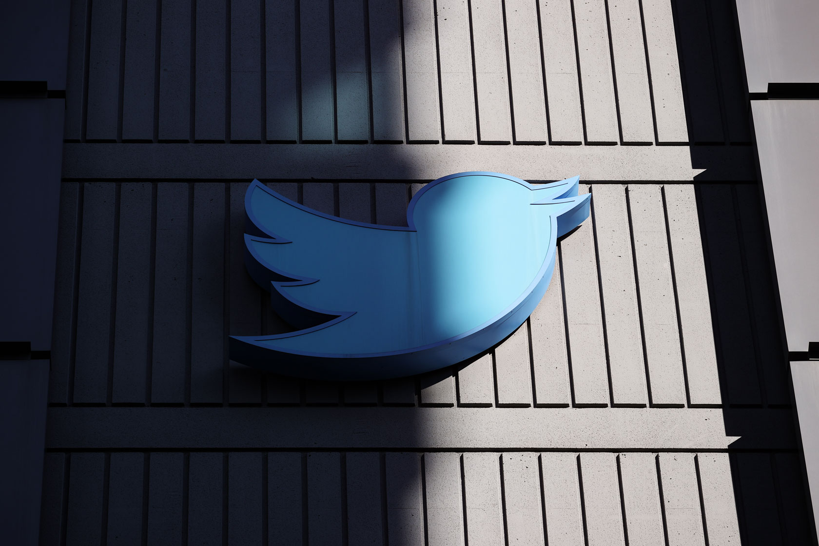 Photo shows the blue Twitter bird logo on the side of a building, partly covered by shade.