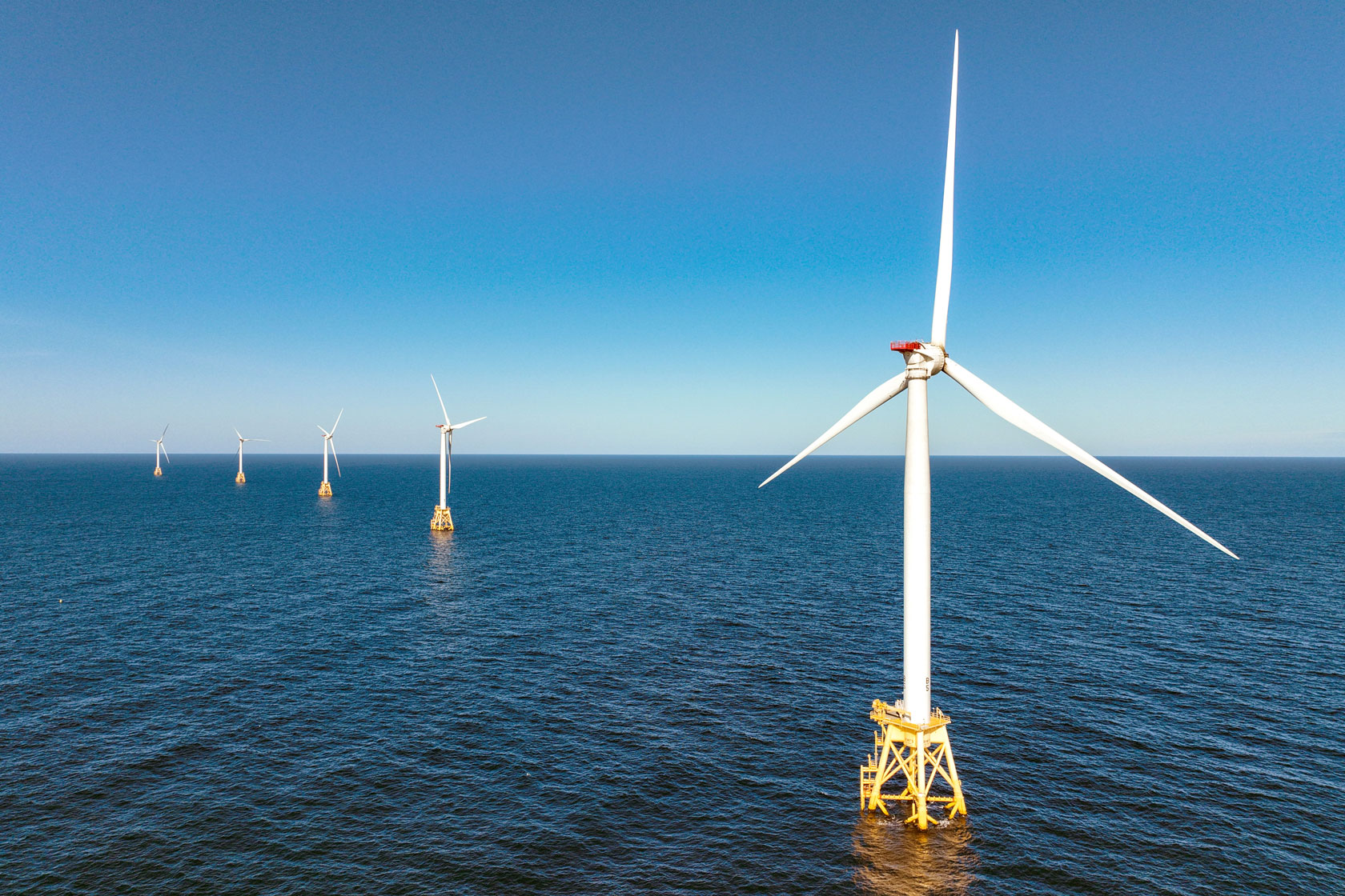 Photo shows a row of five wind turbines sitting in the water on a sunny day.