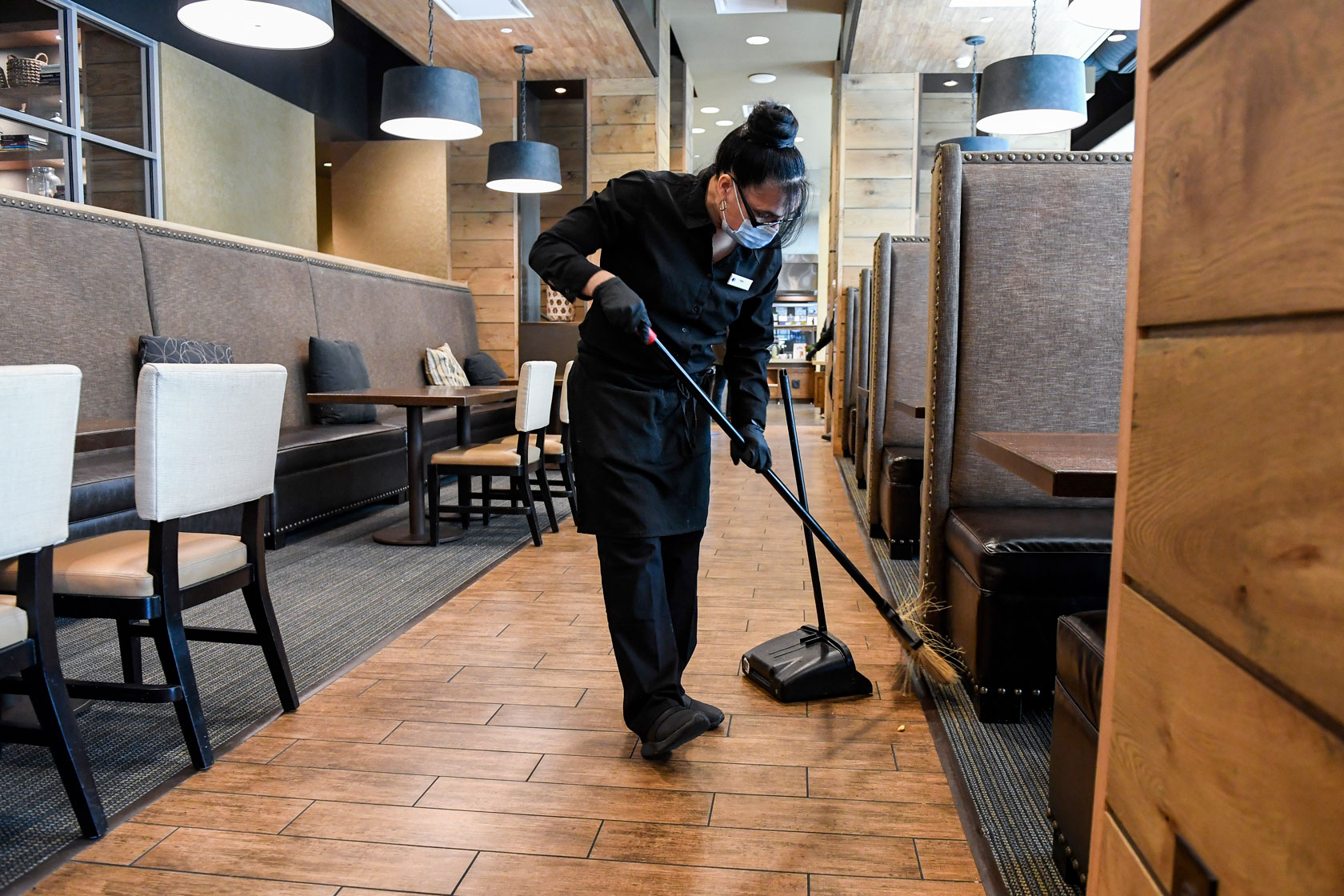 Photo shows a woman in a black uniform sweeping the floor under a booth table in a restaurant.