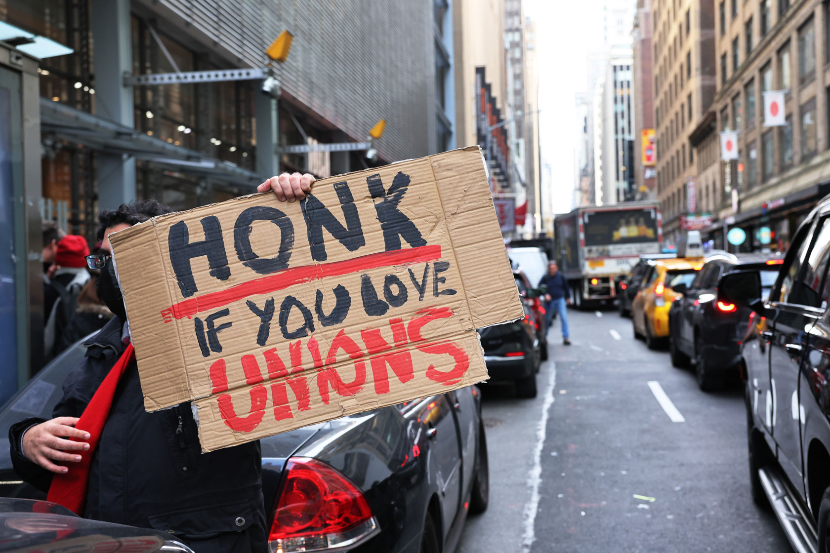 Photo shows a protestor next to cars in the street. The protestor is holding a cardboard sign that says 